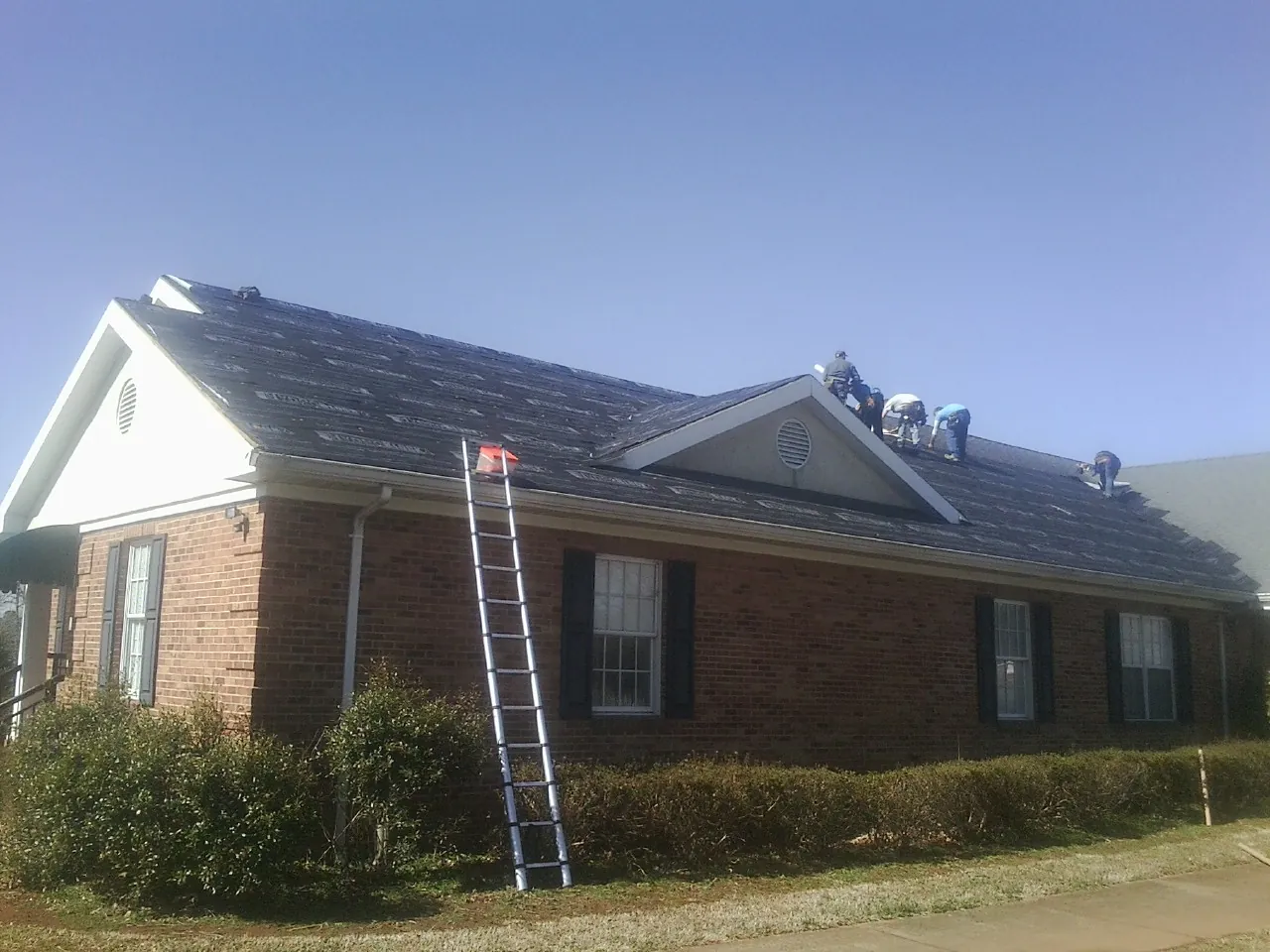 Crew tearing shingles off a brick ranch roof with a ladder set — Metal Roof Repair work by our crew in Hart