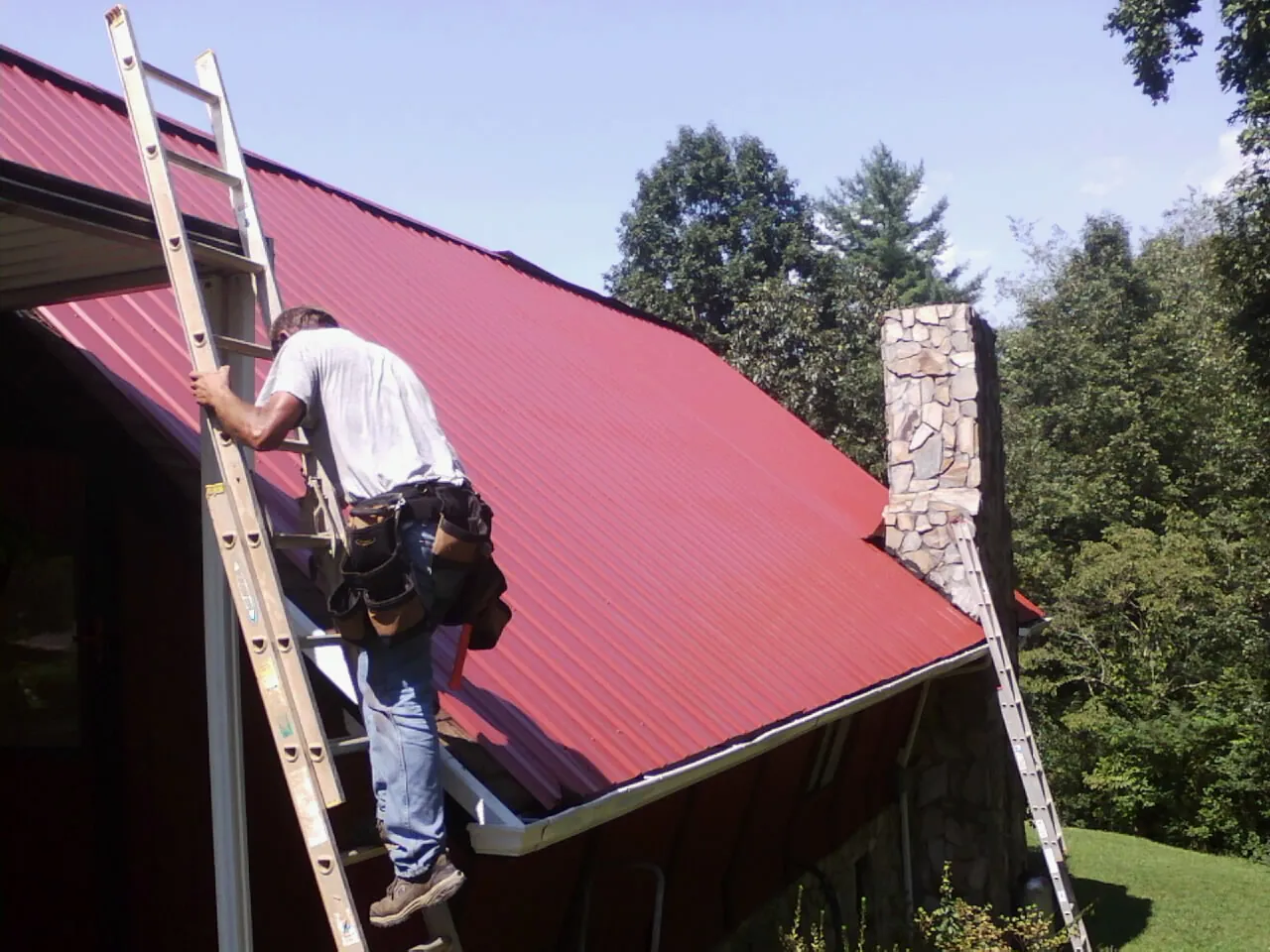 Roofer on a ladder beside a red metal roof and stone chimney — Soffit & Fascia Repair by Roofing Brothers in Hart